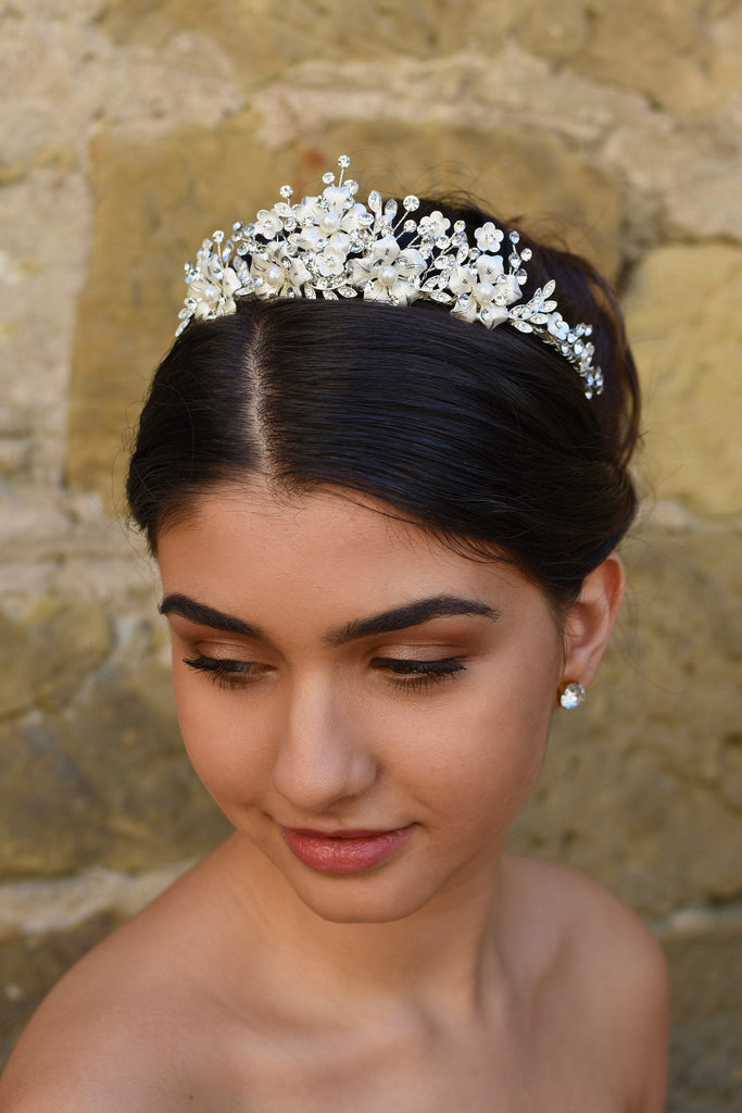 A dark haired model wears a silver tiara with pearl flowers against a stone wall