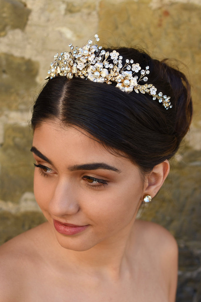 A dark haired model wears a gold tiara with pearl flowers against a stone wall background.