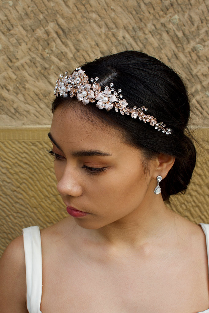 A dark haired model wears a rose gold tiara with pearl flowers against a stone wall background.