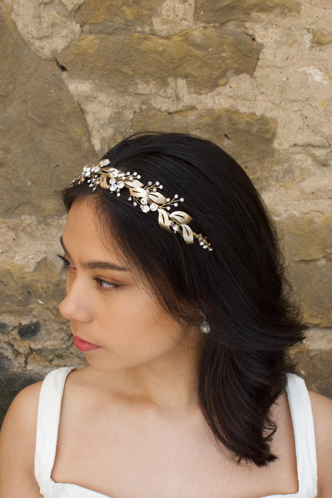 From a side view a Bridal Model with long black hair wears a gold headband of leaf shapes.