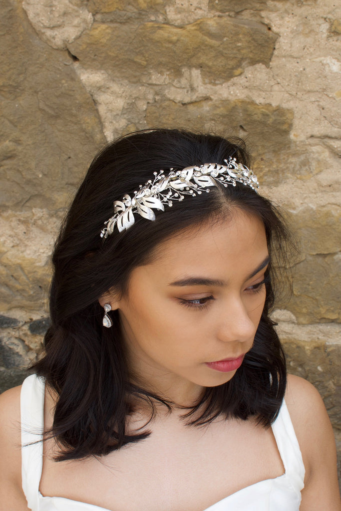 A silver bridal headband is worn by a black haired model with a stone wall in the background
