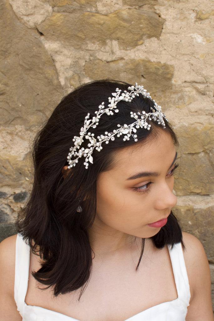 Black hair model looks to the side wearing a silver pearl handmade headband with a stone wall behind