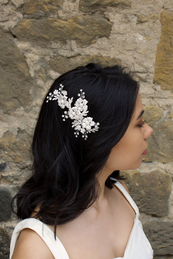A Dark haired model wears a silver side comb of flowers with a stone wall background