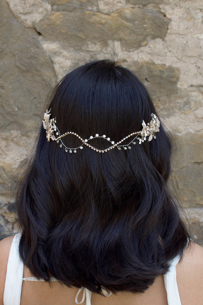 A Black haired model with her hair down wears a soft bridal vine at the back of her head. Stone wall background