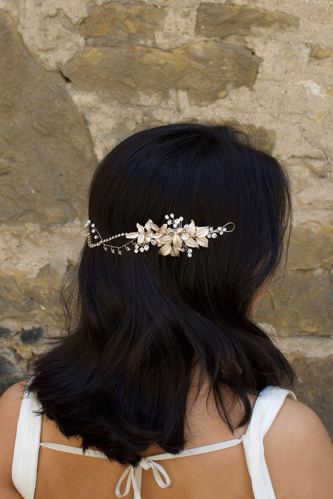 A Black haired model with her hair down wears a soft gold bridal vine at the back of her head. Stone wall background