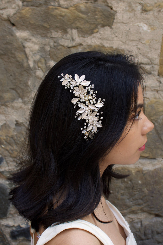 Dark haired model with long hair wears a side comb of pale gold leaves at the side of her head. With a stone wall background