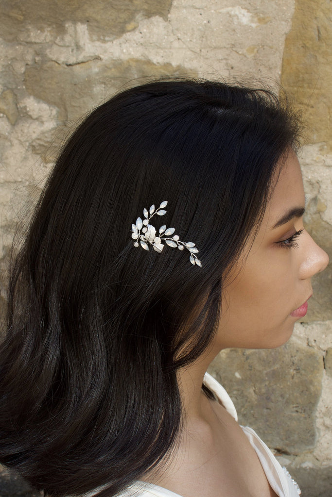 A dark haired model with long hair wears a small leaves bridal hair pin. In the background is a stone wall.