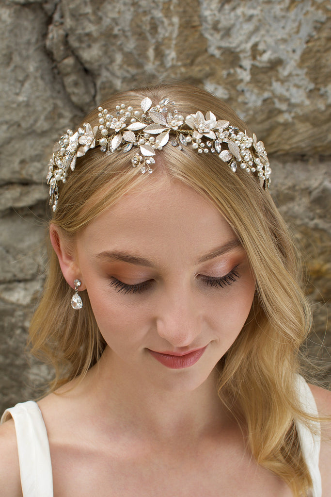 A model Bride with blonde hair wears a gold bridal crown at the front of her head with a stone wall behind