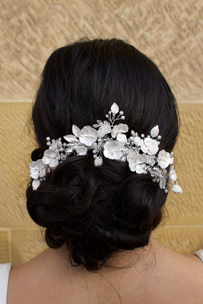Silver flowers and leaves bridal hair piece with a white look worn by a curly haired model with a stone wall background.