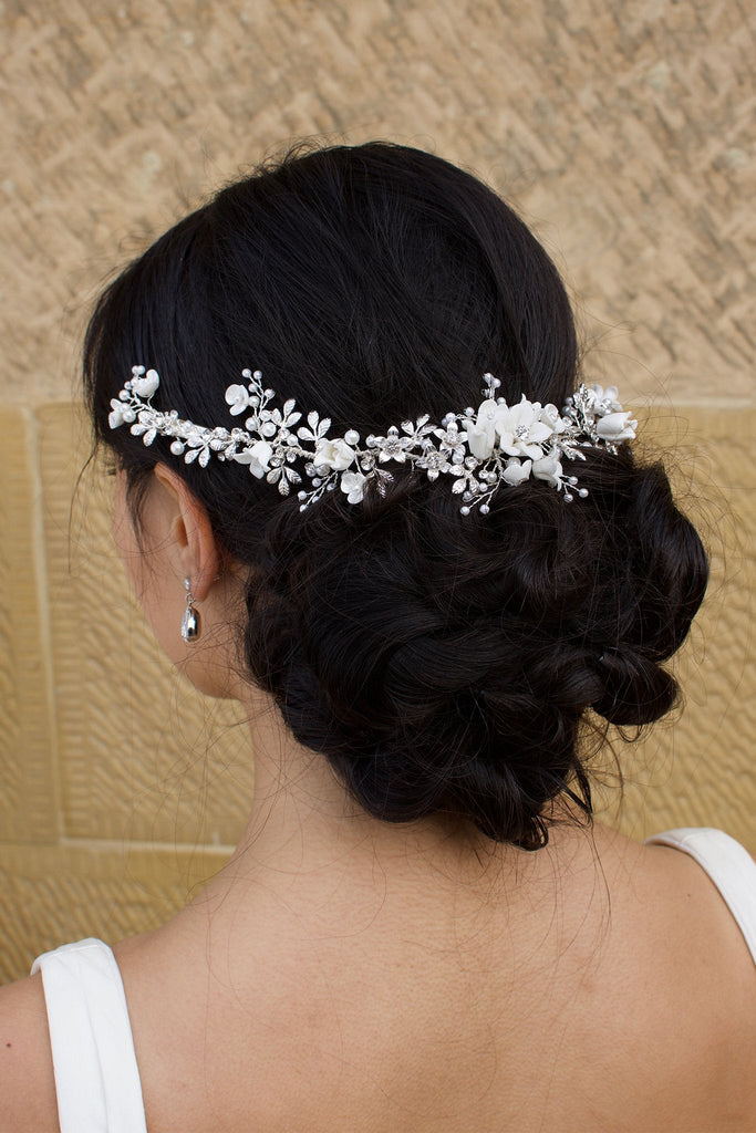 A black haired Bride wears a porcelain flowers silver vine around the back of her head with a stone wall backdrop