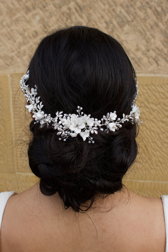 A black haired Bride wears a porcelain flowers silver vine around the back of her head with a stone wall behind