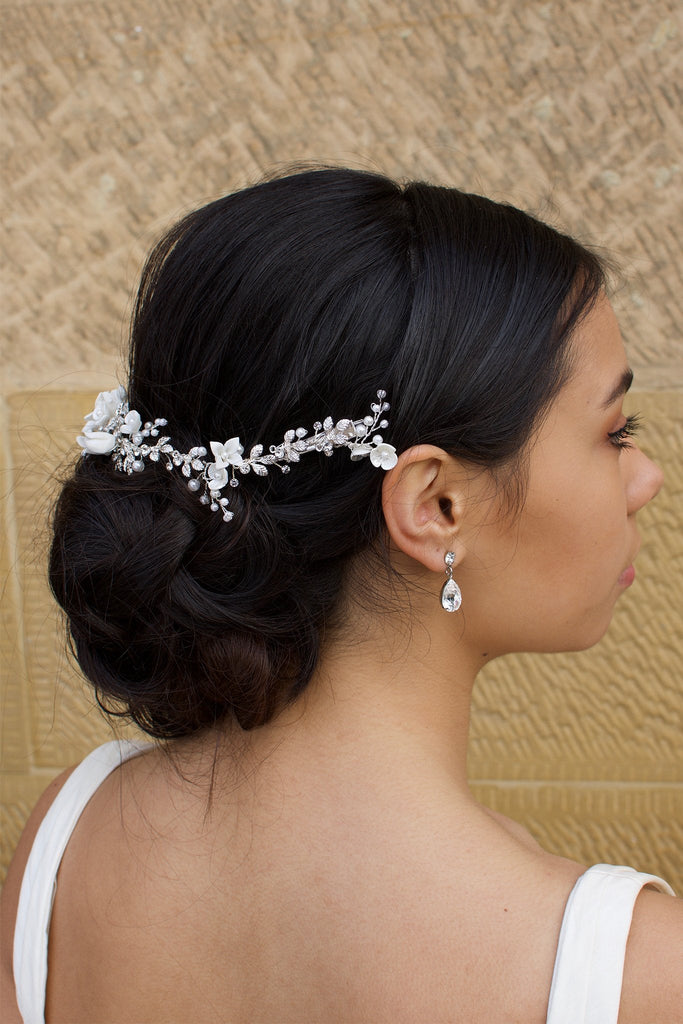 Side view of a black haired Bride wears a porcelain flowers silver vine around the back of her head with a stone wall backdrop