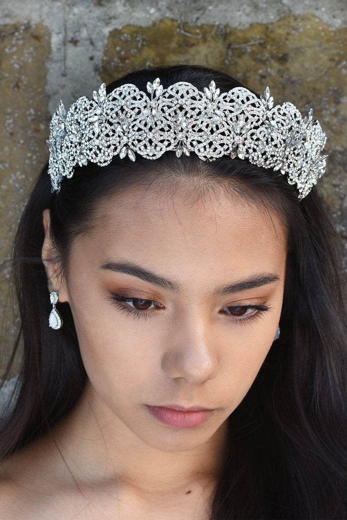 A wide crystal studded tiara is worn at the front of the head of a dark hair model. 